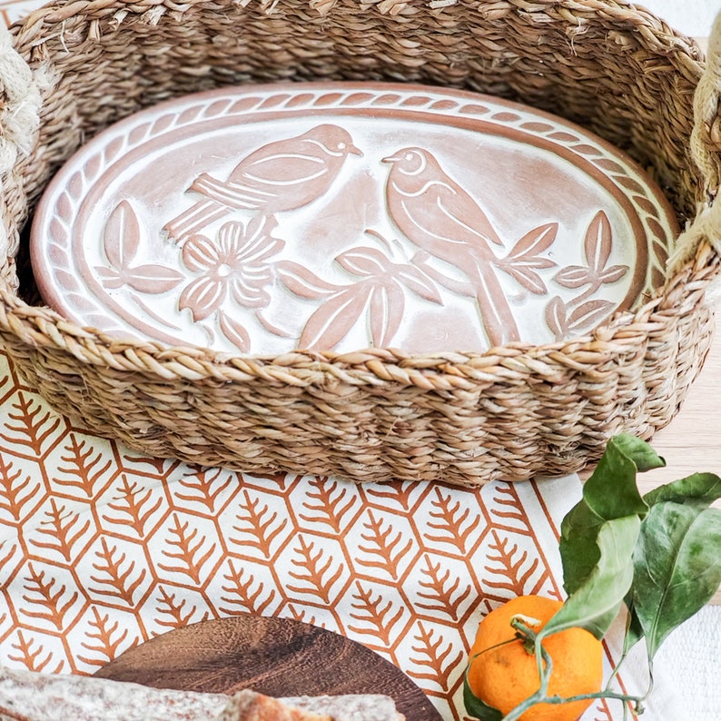 May include: A white ceramic oval serving dish with a raised design of two birds perched on branches with a flower. The dish is sitting inside a woven wicker basket.