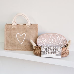 May include: A brown burlap tote bag with a white heart design. Next to it is a wicker basket with a brown and white patterned tea towel and a terracotta oval dish with a white dove design.