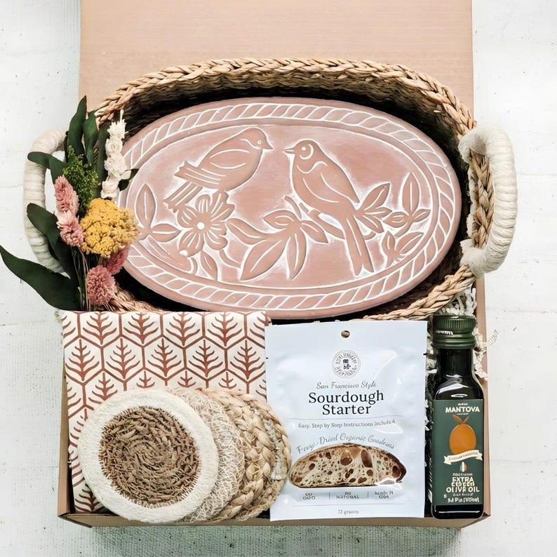 May include: A gift basket containing an oval terracotta tray with a bird and floral relief, a woven seagrass basket, a bundle of dried flowers, a patterned cloth, four round woven coasters, a sourdough starter packet, and a bottle of olive oil.