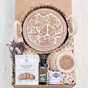 May include: A gift basket with a terracotta serving plate featuring a bird design, a sourdough starter packet, olive oil, coasters, and sea salt. The basket has woven handles and is filled with dried flowers and paper filler.