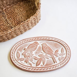 May include: A terracotta oval dish with a raised design of two birds perched on branches with leaves and flowers. The dish has a white glaze and a rope-like border.