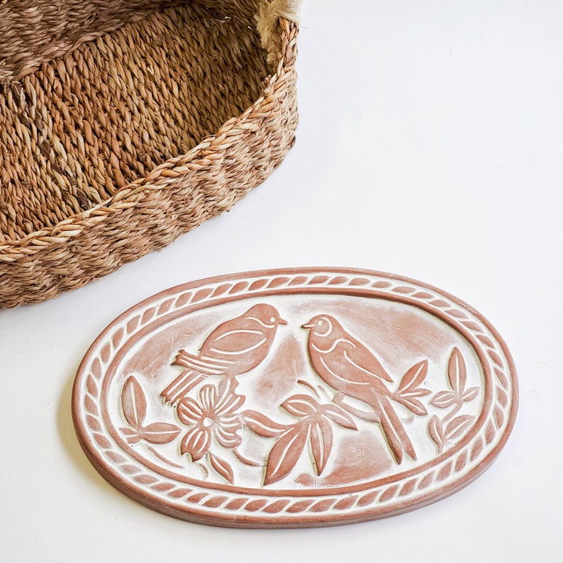 May include: A terracotta oval dish with a raised design of two birds perched on branches with leaves and flowers. The dish has a white glaze and a rope-like border.