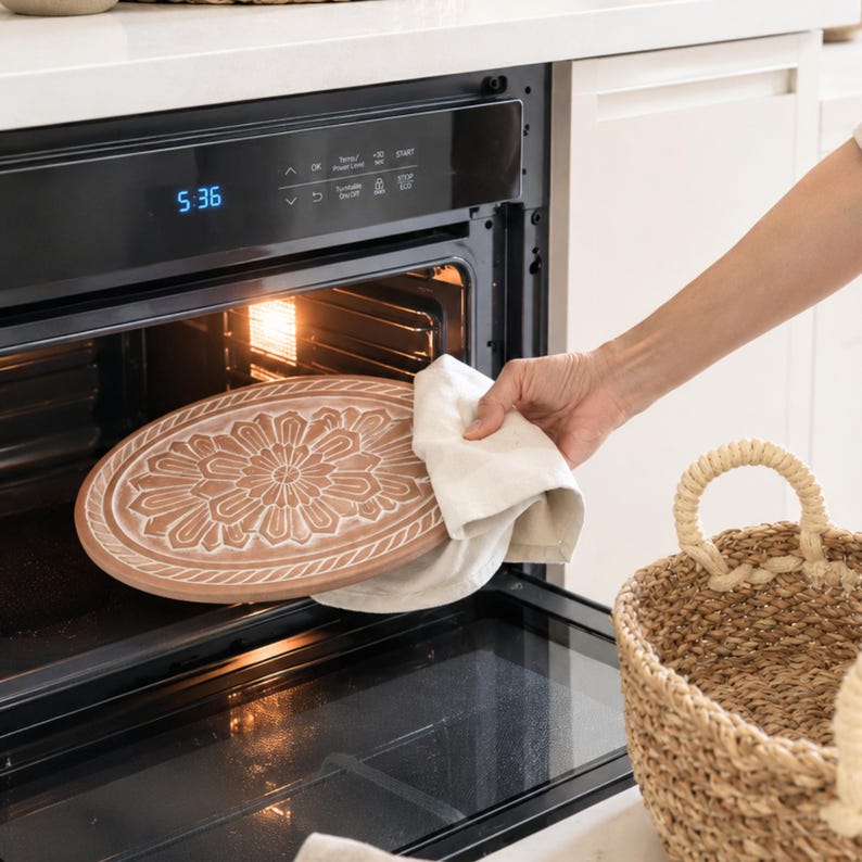May include: A round, light brown wooden tray with a carved floral pattern and a rope-textured border, being held with a white cloth inside a black oven. The oven control panel displays the time 5:36 and various touch-sensitive function icons.