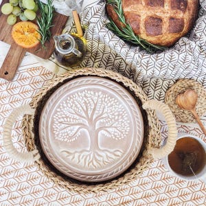 May include: A beige serving tray with a tree of life design, set inside a woven basket. The tray has rope handles. The image also shows a loaf of bread, fruit, olive oil, and tea, suggesting a picnic or dining theme.