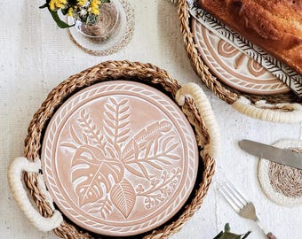 May include: A round terracotta plate with a white embossed botanical leaf pattern, nestled inside a woven seagrass basket with cream-colored rope handles. The plate features detailed monstera and fern leaf designs, providing a natural aesthetic.