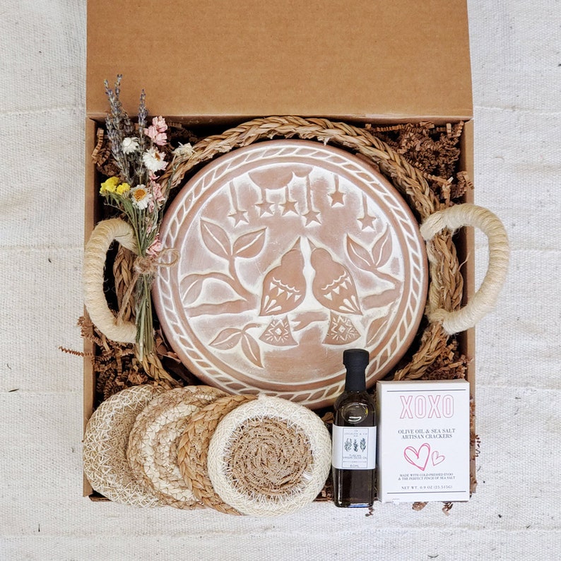 May include: A gift box filled with a terracotta serving dish with a bird design, a bottle of olive oil, woven coasters, and a bag of olive oil and sea salt crackers. The box is lined with brown paper and straw.