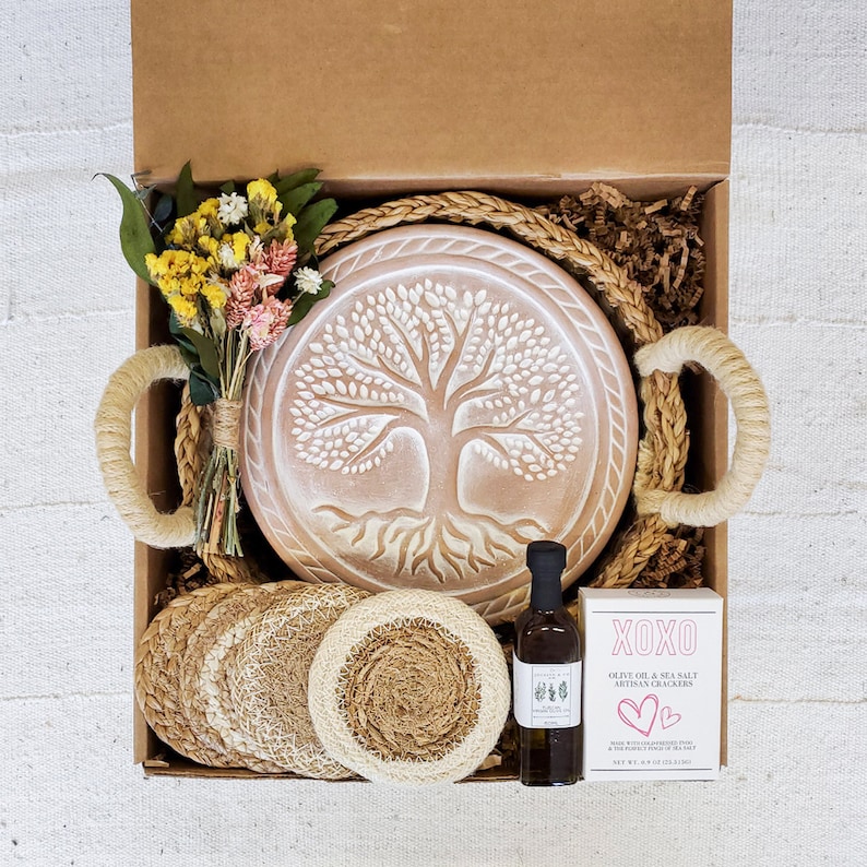 May include: A gift box filled with a variety of items, including a wooden tree of life serving tray, woven coasters, a bottle of olive oil, and a bag of olive oil and sea salt artisan crackers. The box is lined with brown paper and straw.