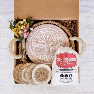 Puede incluir: Una caja de regalo llena de una variedad de art&iacute;culos, que incluyen una base redonda de cer&aacute;mica con un &aacute;rbol de la vida, una bolsa de mezcla para pan de masa madre y posavasos tejidos naturales. La caja est&aacute; forrada con papel marr&oacute;n y paja.