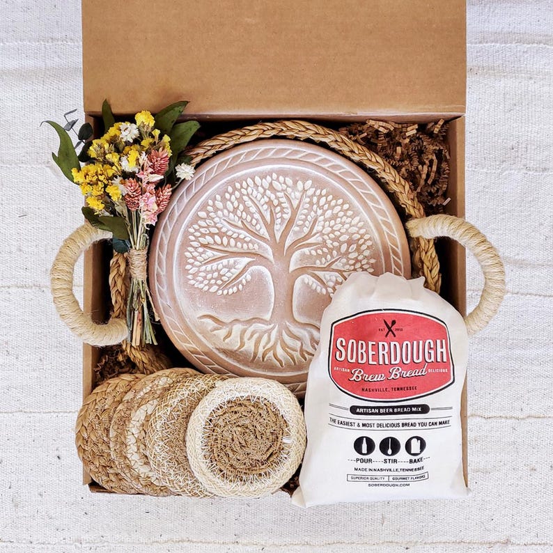 Puede incluir: Una caja de regalo llena de una variedad de art&iacute;culos, que incluyen una base redonda de cer&aacute;mica con un &aacute;rbol de la vida, una bolsa de mezcla para pan de masa madre y posavasos tejidos naturales. La caja est&aacute; forrada con papel marr&oacute;n y paja.
