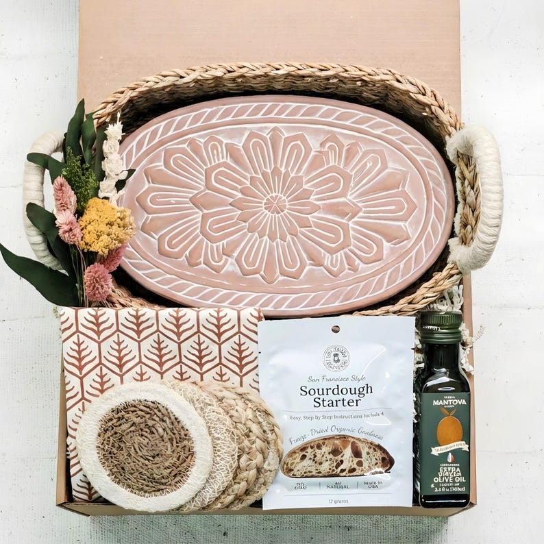 May include: A gift basket containing an oval pink ceramic tray with a floral relief pattern, a small bouquet of dried flowers, a patterned cloth, four round woven coasters, a packet of sourdough starter, and a bottle of Mantova extra virgin olive oil.