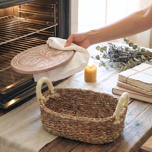 May include: An oval woven seagrass basket with two thick, cream-colored rope handles. The basket features a natural brown texture and a sturdy, open-top design. It is positioned on a wooden surface near a stack of books and a lit candle.