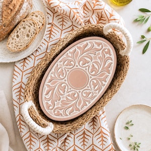 May include: An oval terracotta serving tray featuring a white embossed floral scroll pattern and a central circle, nestled inside a woven seagrass basket with two white rope handles, positioned on a white cloth with an orange leaf print pattern.