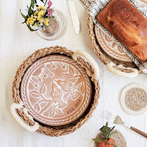 May include: A brown ceramic plate with a white design of two birds and stars. The plate is in a woven basket with two handles. The plate has the text "EST. 2014" on it.