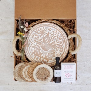 May include: A gift box filled with a brown ceramic plate with a white dove design, a bottle of olive oil and sea salt, and four woven coasters. The box is lined with brown paper and straw.