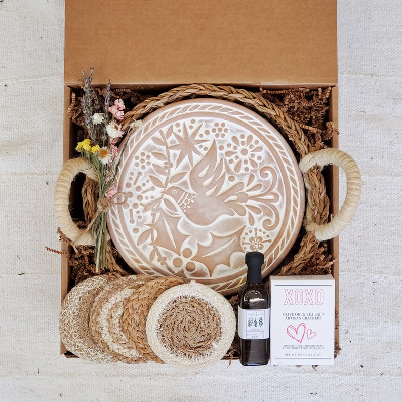 May include: A gift box filled with a brown ceramic plate with a white dove design, a bottle of olive oil and sea salt, and four woven coasters. The box is lined with brown paper and straw.