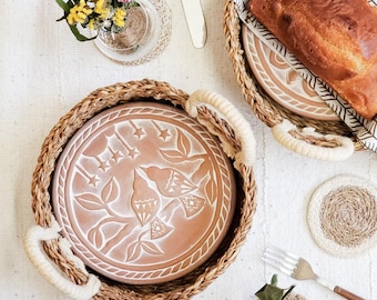 May include: A round terracotta plate featuring an etched design of two birds perched on a branch with stars above, nestled in a woven seagrass basket with cream-colored rope handles. The plate is surrounded by a loaf of bread and a small vase.