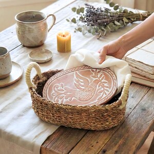 May include: An oval woven seagrass basket with two cream-colored rope handles containing a terracotta-colored ceramic plate featuring an embossed white bird design holding a leafy branch, partially covered by a folded white cloth napkin.