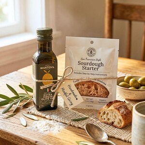 May include: A glass bottle of Mantova Italian Golden extra virgin olive oil, 3.4 fl oz, tied with twine to a white pouch of San Francisco Style Sourdough Starter, 1 oz, featuring a small tag labeled Handmade & Mindful Giving, with a piece of bread.
