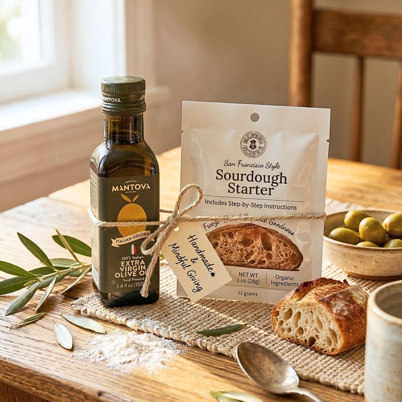 May include: A glass bottle of Mantova Italian Golden extra virgin olive oil, 3.4 fl oz, tied with twine to a white pouch of San Francisco Style Sourdough Starter, 1 oz, featuring a small tag labeled Handmade & Mindful Giving, with a piece of bread.