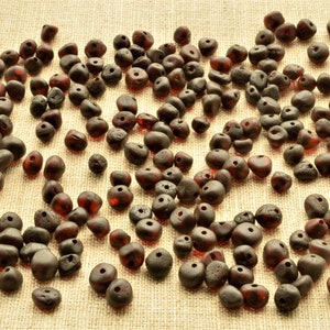 May include: A large collection of dark red amber beads, each with a hole drilled through the centre. The beads are scattered on a light brown fabric surface.