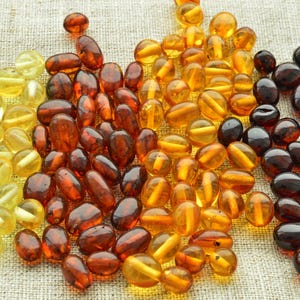 May include: A selection of amber beads in shades of yellow, orange, and dark brown. The oval-shaped beads have a polished, glossy finish. The beads are arranged on a neutral-toned, textured surface, creating a visually appealing display.