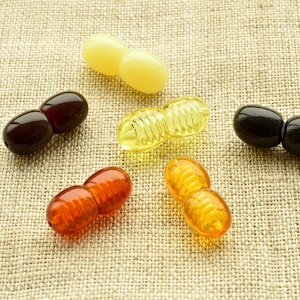 May include: Six pairs of amber beads, each pair connected together. The beads are in various shades of amber, including yellow, orange, and brown. The beads are arranged in a semi-circle on a beige fabric background.