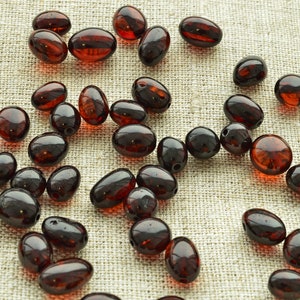 May include: A scattering of dark amber beads in various shapes. The beads are oval, round, and irregular, with a glossy, transparent appearance. They are scattered on a light tan, textured surface, suggesting a craft or jewellery-making theme.