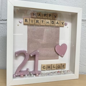 May include: A white picture frame with a pink glitter background. The frame contains the words "HAPPY BIRTHDAY" spelled out with wooden letter tiles, the number "21" in pink, a pink heart, and the name "CHLOE" spelled out with wooden letter tiles. The frame is decorated with white and pink pearls.