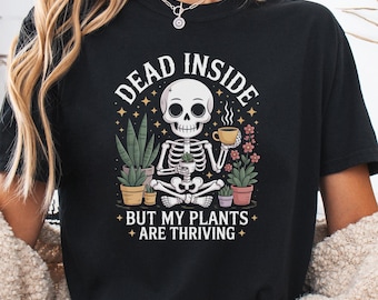 Pode incluir: Camiseta preta com um gráfico apresentando um esqueleto, plantas e o texto "DEAD INSIDE BUT MY PLANTS ARE THRIVING". O design inclui um esqueleto segurando uma xícara, cercado por plantas em vasos. A camiseta é de estilo casual.