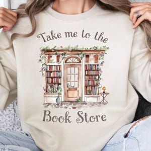 May include: Sand-colored sweatshirt with the text "Take me to the Book Store" above a watercolor illustration of a bookstore facade. The design features bookshelves, a door, and floral accents. The sweatshirt is a neutral color.