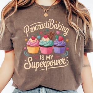 May include: Espresso-colored t-shirt featuring the phrase "ProcrastiBaking is my Superpower" with three cupcakes. Each cupcake has a different frosting color and topping, including cherries, chocolate, and strawberries. The shirt is a casual, comfortable style.
