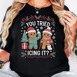 May include: Black t-shirt with a festive gingerbread man design. The shirt features the text "YOU TRIED ICING IT?" above two gingerbread men, one in scrubs and the other with a leg cast. Christmas tree and snowflake graphics complete the design.