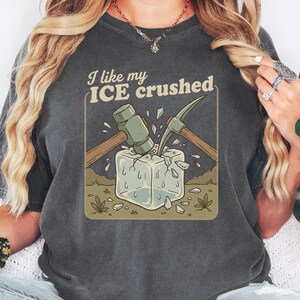 May include: Dark grey t-shirt with a graphic of a hammer and pickaxe breaking ice, with the text "I like my ICE crushed". The shirt is a relaxed fit and the colour is described as pepper.