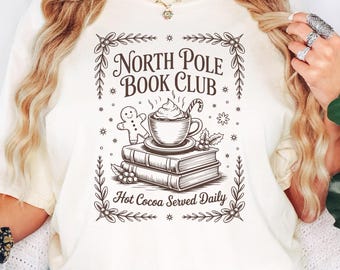 Op de afbeelding: Ivoor t-shirt met een afbeelding van "North Pole Book Club". Het ontwerp toont een kop warme chocolademelk, een peperkoekmannetje en boeken. De zin "Hot Cocoa Served Daily" is ook inbegrepen.