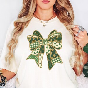 May include: Ivory-colored t-shirt featuring a large, patchwork bow design in shades of green and gold. The bow incorporates various patterns, including shamrocks and plaid. The shirt is a comfortable, casual style.