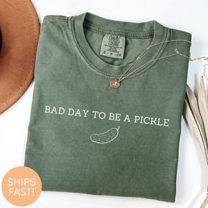 Puede incluir: Camiseta verde oliva con la frase blanca "BAD DAY TO BE A PICKLE" y una ilustración de pepinillo. La camiseta tiene cuello redondo y mangas cortas. Un collar dorado con un colgante está sobre la camiseta.
