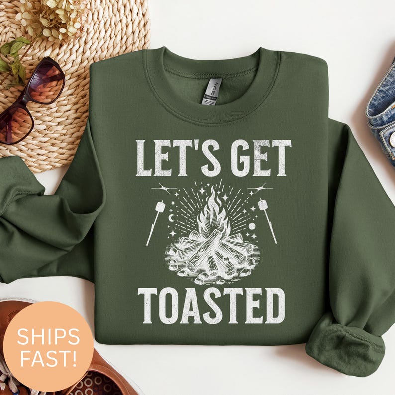 Op de afbeelding: Groen sweatshirt met de tekst "LET'S GET TOASTED" boven een afbeelding van een kampvuur met marshmallows. Het sweatshirt is effen groen van kleur.