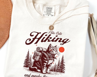 May include: A beige t-shirt with a red graphic of a cat wearing a backpack and hiking in the woods. The text on the shirt reads "I like Cats Hiking and maybe three people".