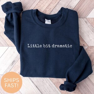 Könnte beinhalten: Marineblaues Sweatshirt mit dem weißen Schriftzug "Little bit dramatic". Das Sweatshirt ist gefaltet und liegt auf einer hellen Holzmaserung. Die Ärmel sind umgeschlagen.