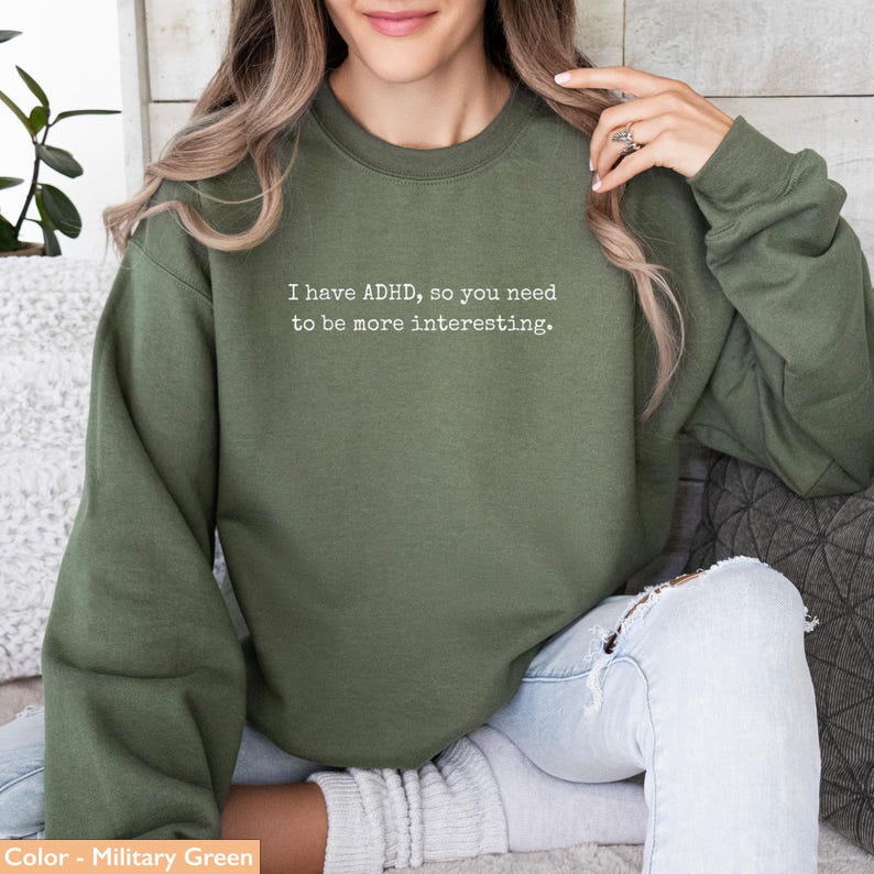 May include: Military green sweatshirt with the text "I have ADHD, so you need to be more interesting." The long-sleeved top is a solid color and has a crew neck.