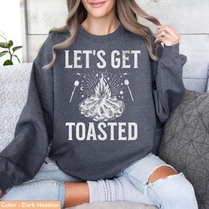 Op de afbeelding: Donkergrijze sweatshirt met de witte tekst "LET'S GET TOASTED". Het ontwerp toont een kampvuur en marshmallows op stokjes. Een comfortabele top voor kamperen of buitenactiviteiten.