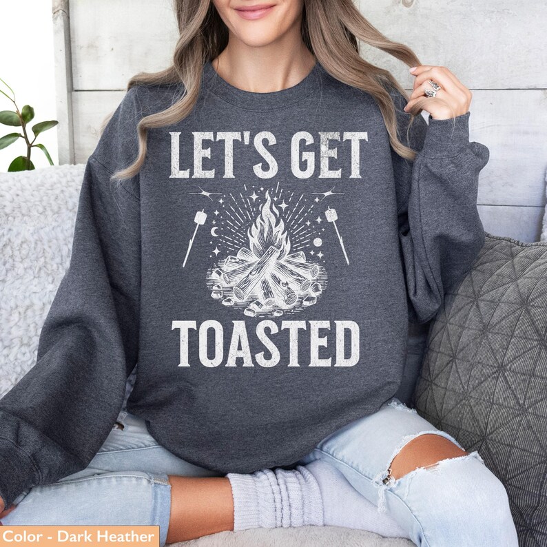 Op de afbeelding: Donkergrijze sweatshirt met de witte tekst "LET'S GET TOASTED". Het ontwerp toont een kampvuur en marshmallows op stokjes. Een comfortabele top voor kamperen of buitenactiviteiten.