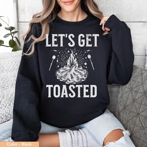 Op de afbeelding: Zwarte sweatshirt met de witte tekst "LET'S GET TOASTED", met een kampvuurafbeelding met marshmallows op stokjes. Het ontwerp bevat een vreugdevuur met vlammen, sterren en maandetails. Een casual, comfortabele top.