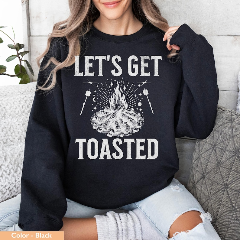 Op de afbeelding: Zwarte sweatshirt met de witte tekst "LET'S GET TOASTED", met een kampvuurafbeelding met marshmallows op stokjes. Het ontwerp bevat een vreugdevuur met vlammen, sterren en maandetails. Een casual, comfortabele top.