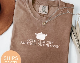 Puede incluir: Una camiseta marrón Comfort Colors con el texto "OOPS, I BOUGHT ANOTHER DUTCH OVEN" y un dibujo de una olla holandesa. Un collar plateado con un colgante está sobre la camiseta. La camiseta es talla S-P-CH.