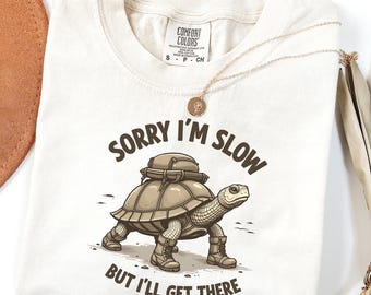 May include: A beige t-shirt with a cartoon illustration of a turtle carrying a backpack. The text "Sorry I'm Slow But I'll Get There" is printed on the shirt.