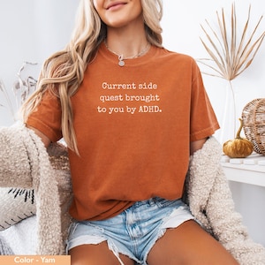 May include: A woman wearing a burnt orange t-shirt with the text "Current side quest brought to you by ADHD." The shirt is made of a soft, comfortable material.