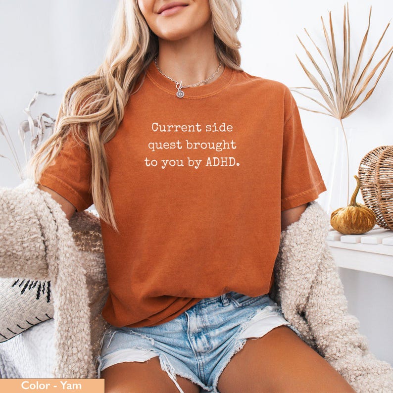 May include: A woman wearing a burnt orange t-shirt with the text "Current side quest brought to you by ADHD." The shirt is made of a soft, comfortable material.