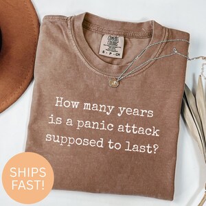 Op de afbeelding: Een bruin T-shirt met witte tekst die luidt: "Hoeveel jaar duurt een paniekaanval?"