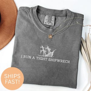 Puede incluir: Una camiseta gris Comfort Colors con el texto "I RUN A TIGHT SHIPWRECK" y un gráfico de barco. Un collar plateado con un colgante está sobre la camiseta. Un sombrero marrón y hojas decorativas están en el fondo.